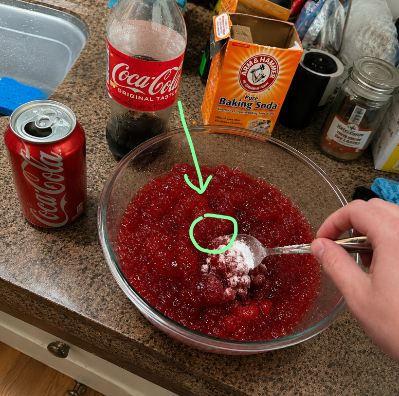 A glass bowl of red gelatin with baking soda on a spoon, Coca-Cola, and pantry ingredients on a kitchen counter
